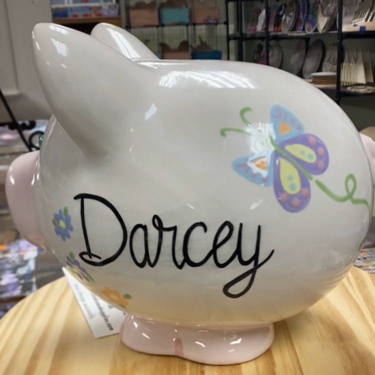 White piggy bank with 'Darcey' written on it, sitting on a wooden surface.